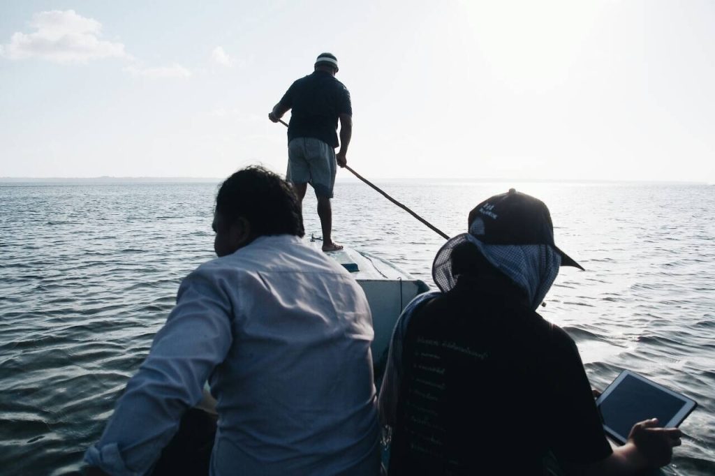 Photo of two people in narrow fishing boat, one is holding and looking at an iPad, the other is looking at the water. The third person is steering the boat at the bow end.